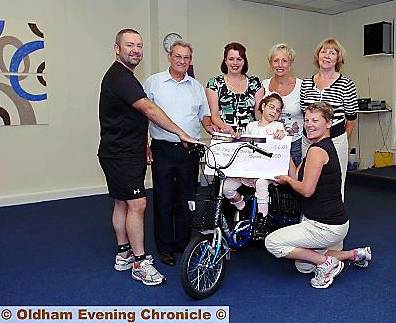 Cordelia Cowsill tries out the tricycle at the cheque presentation, with (from the left) Derek Ireland, co-owner of Lifestyle Fitness Centre, Bill Bussey, Rotary Club of Saddleworth, Amanda Cowsill, fundraising co-ordinators Gill Bussey, and Ann Beswick, and (kneeling) Lynne Furness, gym co-owner 